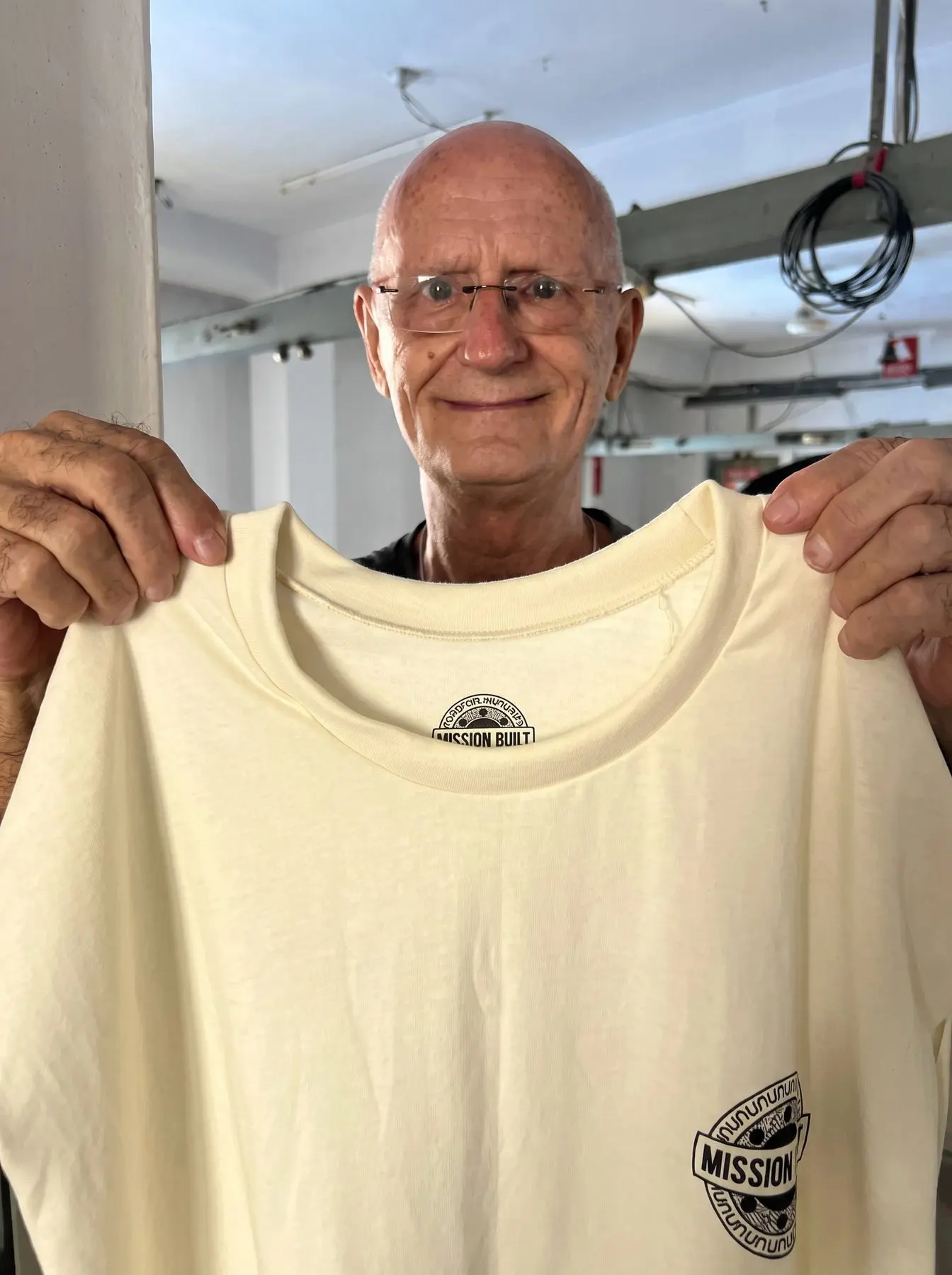 A man in glasses holding up a finished cream t-shirt on the factory floor
