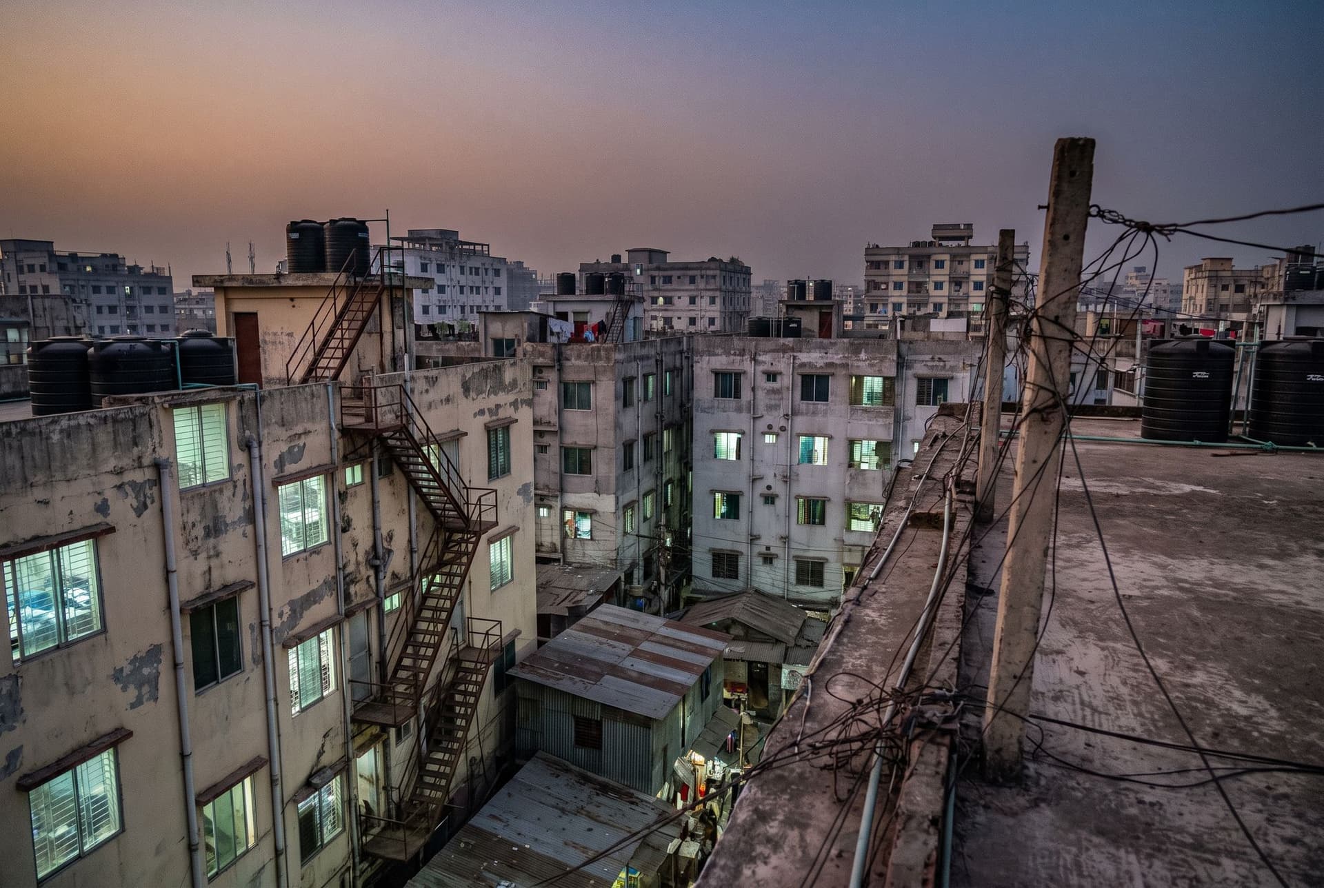 Dusk view from a rooftop over dense multi-storey factory buildings with lit windows, black water tanks and tangled power lines